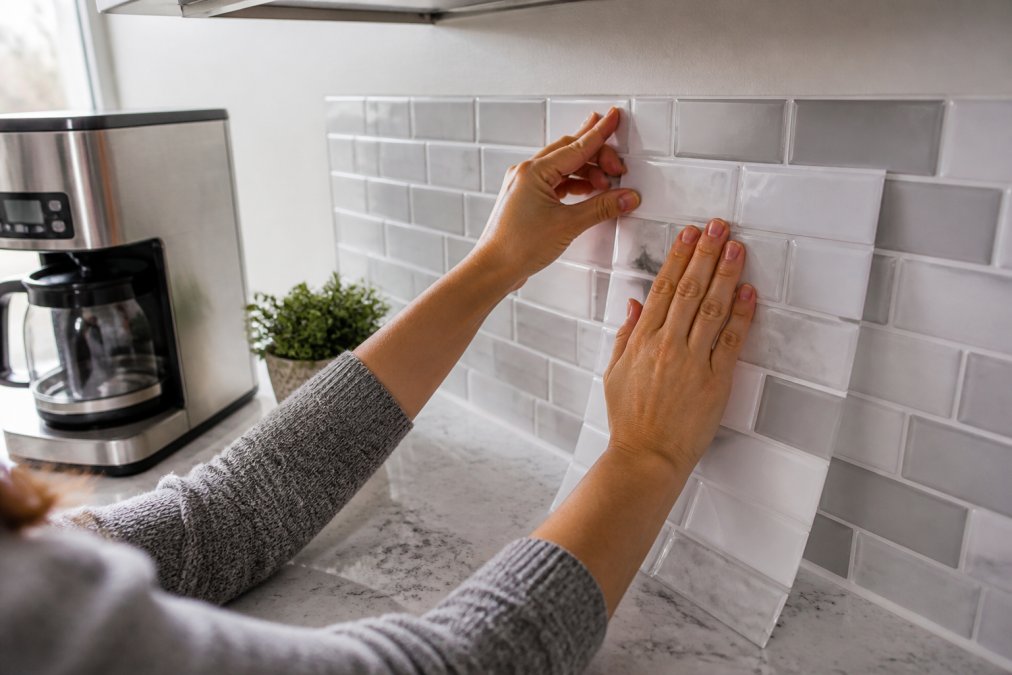 How to Upgrade a Rental Kitchen Without Renovation 2 Close-up of hands pressing a peel-and-stick backsplash tile onto a kitchen wall above a quartz countertop, with a stainless steel coffee maker and a small potted plant nearby in soft natural light