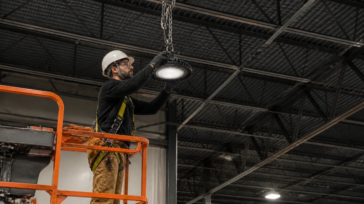 Professional electrician installing high-bay LED fixture in tall warehouse space with safety harness, aerial lift equipment, and proper rigging setup visible