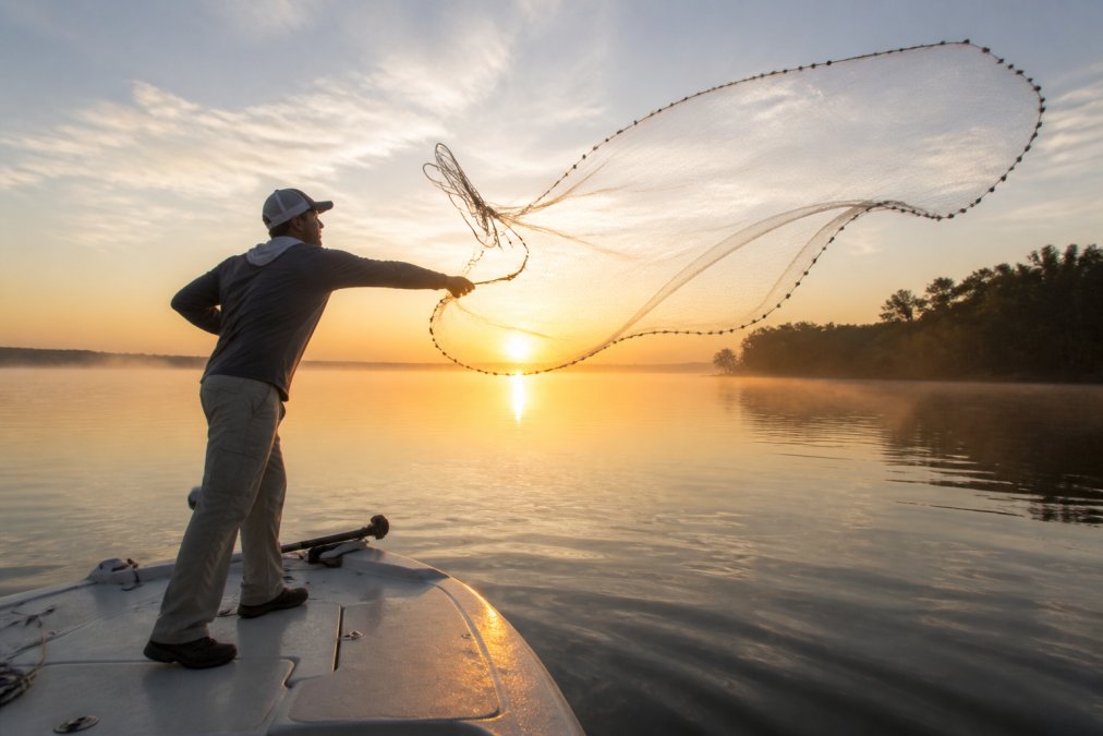 Angler on a boat deck throwing a wide cast net over calm water at sunrise