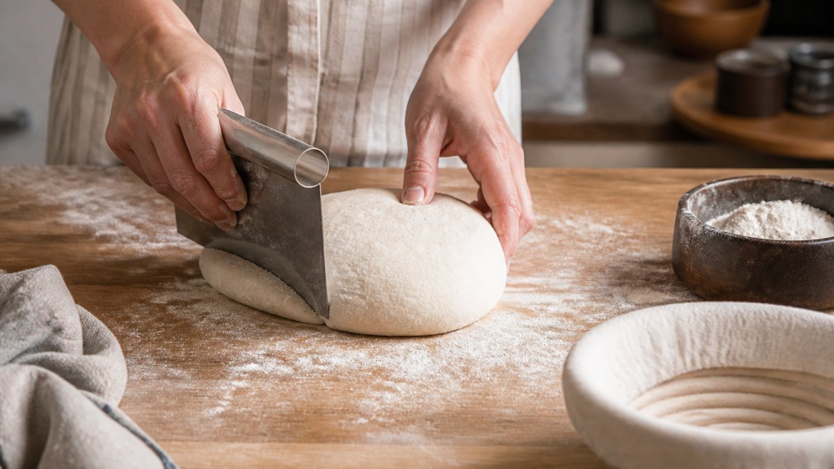 Close-up of two hands using a stainless steel bench scraper to push and rotate a round of sourdough on a floured wooden countertop, dough pulling into a taut ball, soft window light