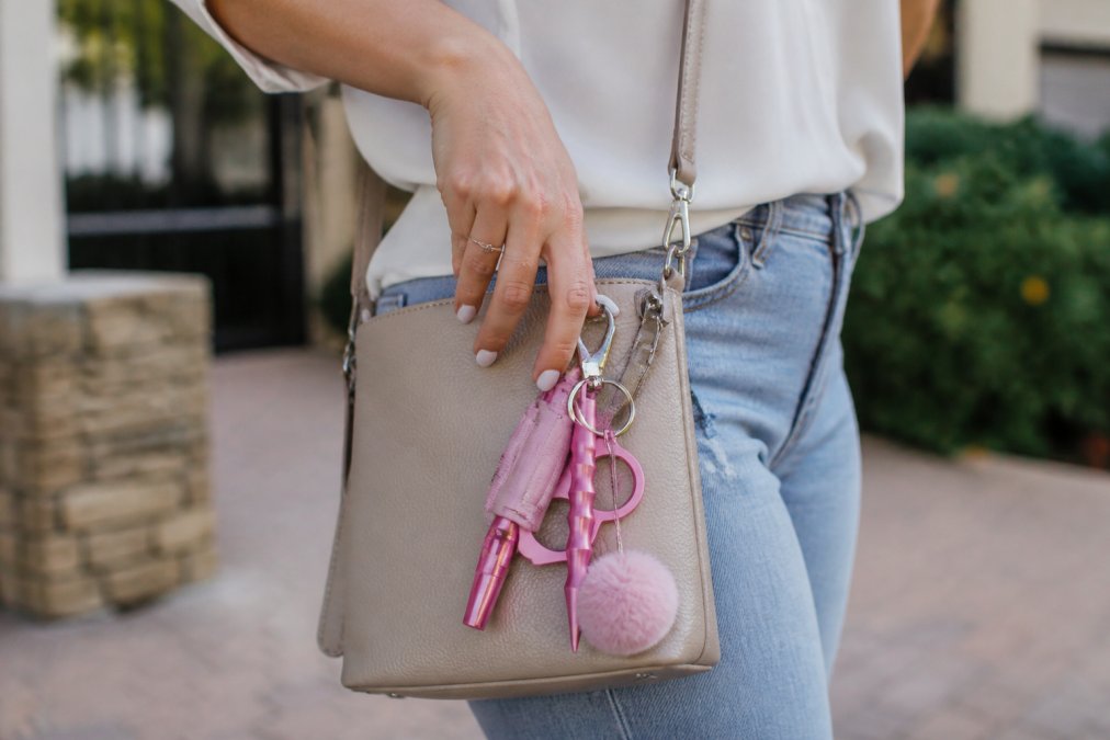 Woman's hand positioned near a stylish safety keychain clipped to the outside of her crossbody bag, demonstrating quick-access placement in natural daylight