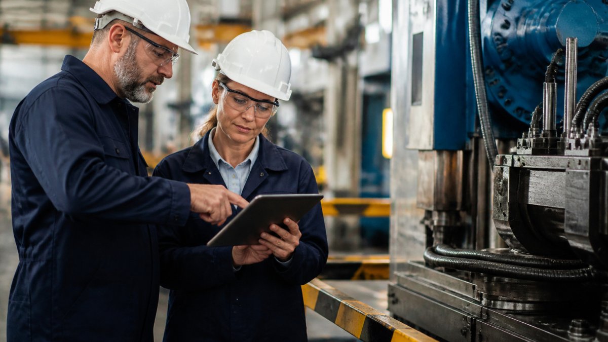 Technician and supervisor using a tablet to review factory documentation logs beside industrial machinery