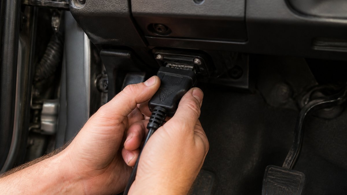 Close-up of a mechanic's hands plugging an OBD-II scanner into the 16-pin diagnostic port under a car's dashboard, ignition on, workshop lighting