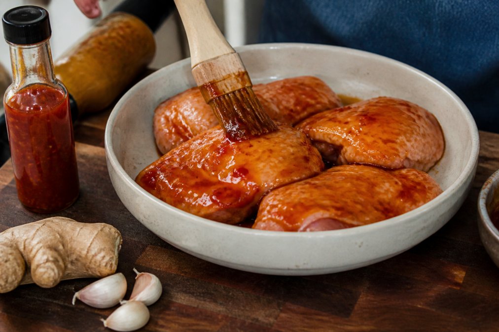 Raw chicken thighs being brushed with glossy chilli jam marinade in a ceramic bowl, with fresh ginger and garlic cloves nearby on a wooden cutting board