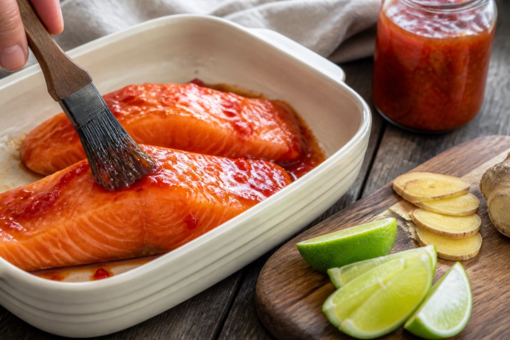 Raw salmon fillets being brushed with glossy chilli jam marinade in a ceramic dish, with fresh lime wedges and ginger slices arranged on a wooden board beside it