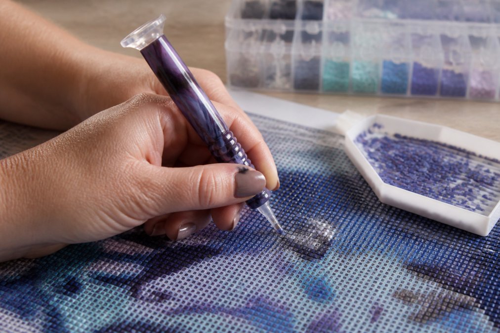 Close-up of hands holding an ergonomic diamond painting pen with correct neutral wrist alignment, stylus positioned at 45-degree angle over canvas with organized drills in grooved tray