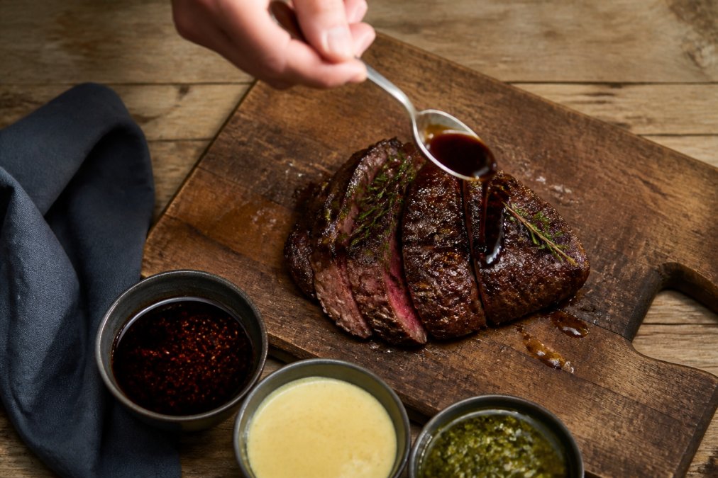 Close-up overhead shot of a hand drizzling a dark artisan finishing sauce over a plated seared steak on a rustic wooden cutting board, with small bowls of different sauces nearby