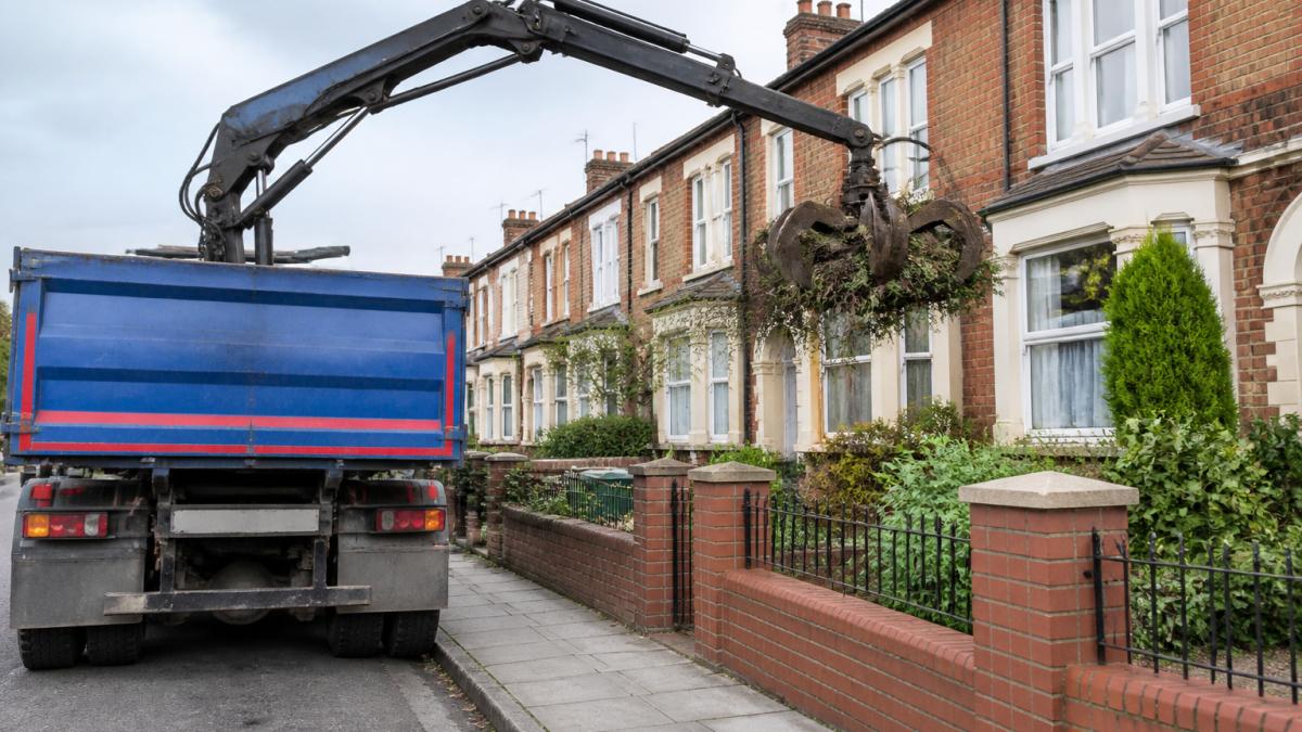 Grab lorry crane reaching over a garden wall to collect green waste for a residential grab hire service.