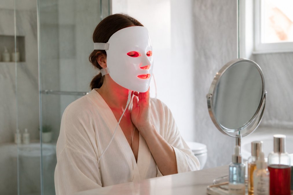 A woman in her 40s sitting at a vanity, wearing a sleek white LED face mask that emits a soft red glow, with a small mirror and skincare bottles nearby in a bright, modern bathroom