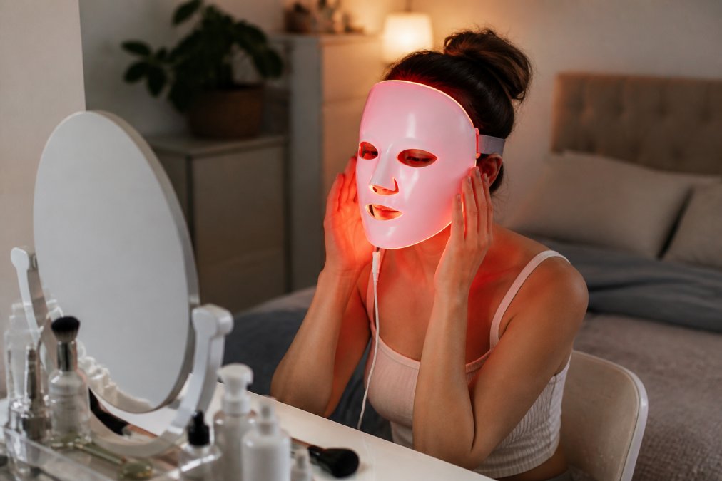 A young woman sitting at a vanity, wearing a flexible red LED face mask, eyes protected with goggles, soft ambient lighting in a modern bedroom