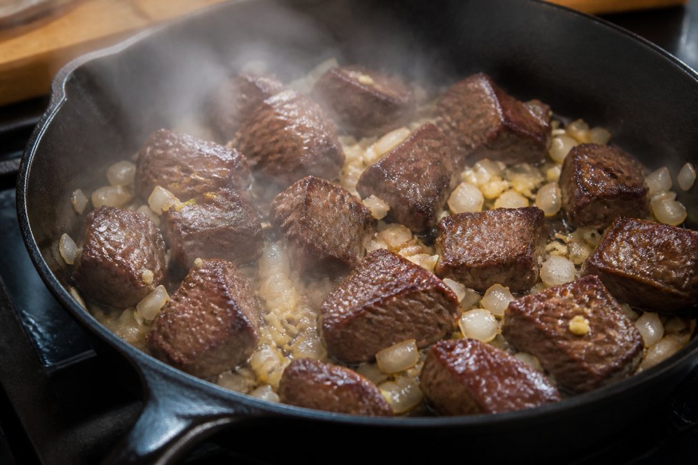 Close-up of seared beef chunks in cast iron skillet with deep brown crust, minced garlic and diced onions scattered around, steam rising from the pan under warm kitchen lighting