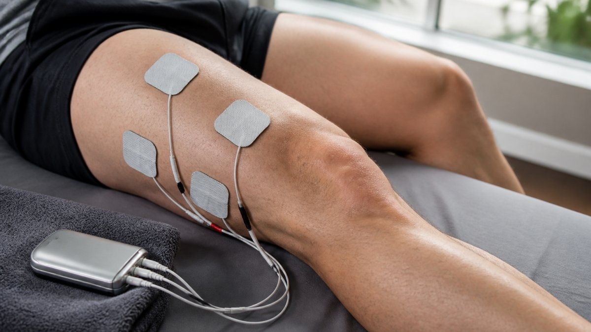 Close-up of a person's leg with electrode pads placed on the quadriceps, portable muscle stimulator unit resting on a towel nearby, soft daylight from a window