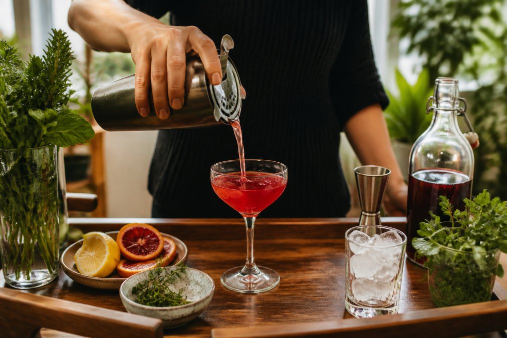 Home bartender pouring a vibrant shrub cocktail into a coupe glass on a wooden bar cart with fresh herbs and citrus