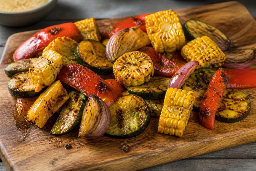 Colorful roasted vegetables including bell peppers, zucchini, corn, and red onions coated in Southwest seasoning on a wooden cutting board, glistening with oil and char marks