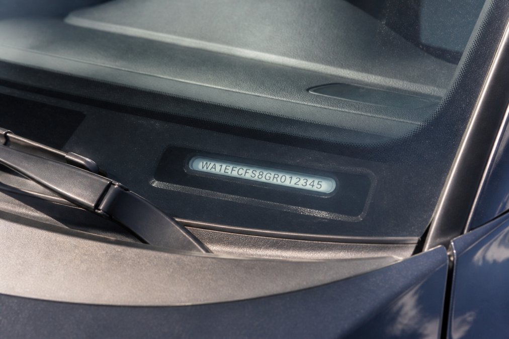 Close-up of a vehicle's dashboard showing the VIN plate visible through the windshield on the driver's side, with sunlight highlighting the 17-character alphanumeric code clearly visible