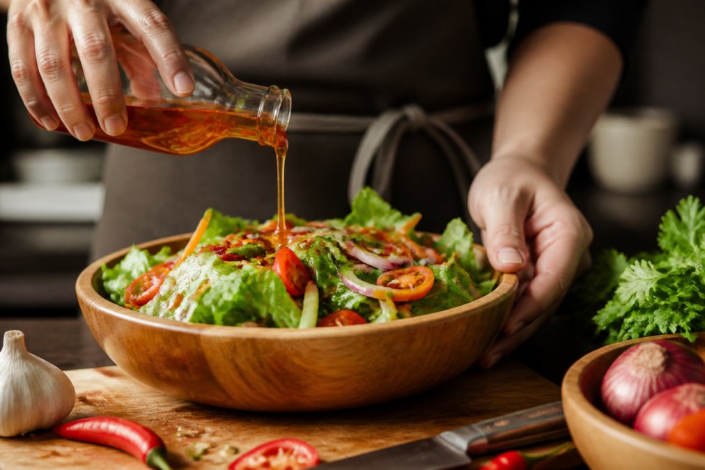 Close-up of hands drizzling chilli vinegar over a vibrant salad in a wooden bowl, with fresh herbs and vegetables visible