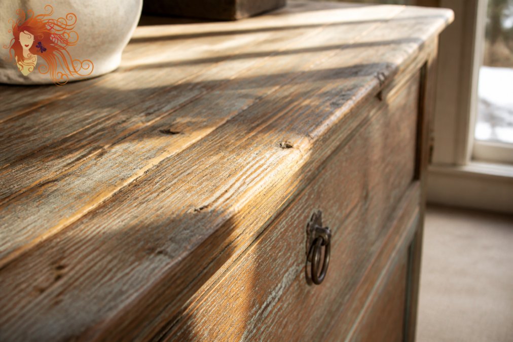 Close-up of distressed vintage wood furniture piece showing weathered texture, natural patina, and visible wood grain details illuminated by natural sunlight streaming through a window