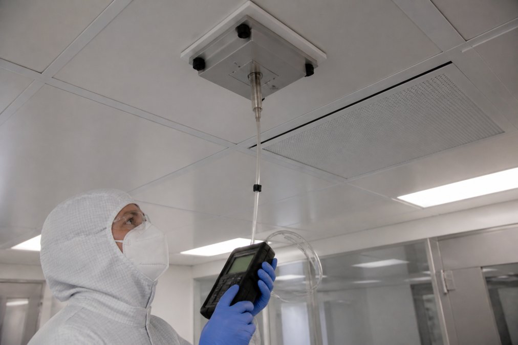 Technician performing particle counting test with calibrated monitoring equipment mounted in cleanroom ceiling, collecting data from HEPA filter validation with digital display visible