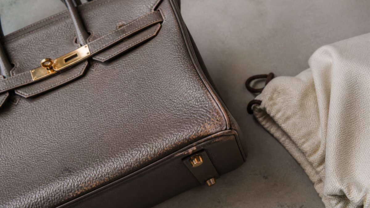 Close-up overhead photo of a structured leather handbag on a neutral surface, showing corner wear, hardware scratches, and a dust bag beside it, shot in soft natural window light