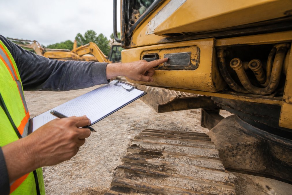Close-up of hands holding equipment inspection clipboard while checking stamped serial number plate on heavy machinery in outdoor equipment yard, natural lighting