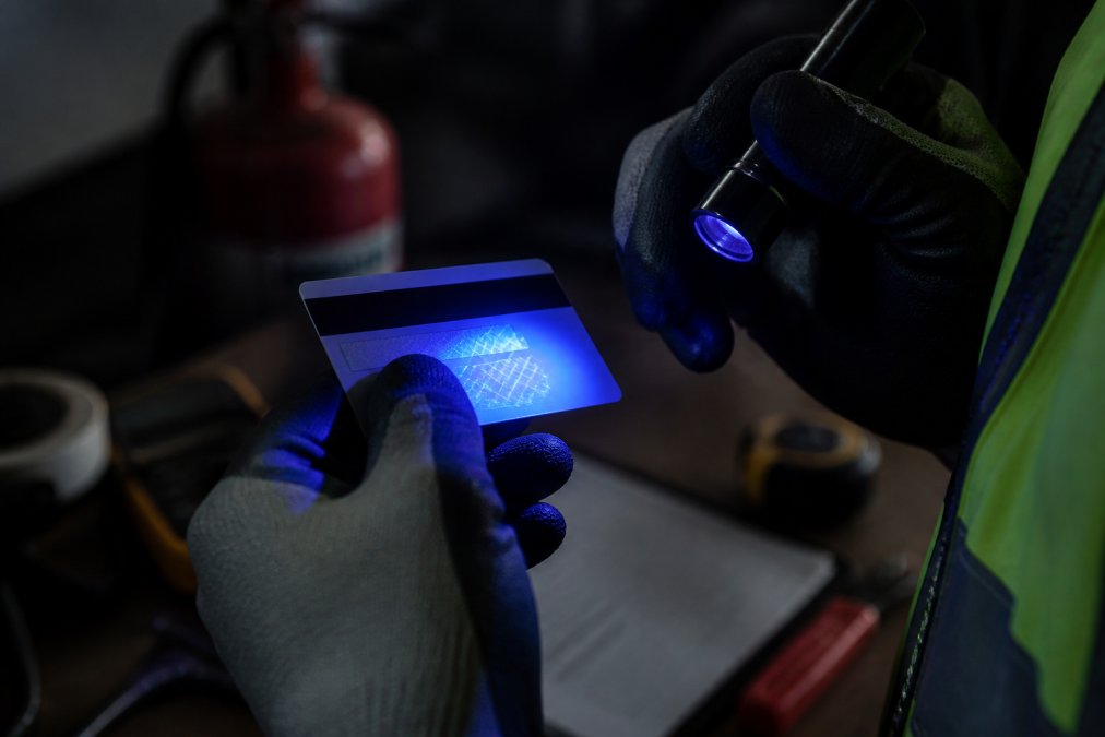 Close-up of a gloved hand holding a fire protection contractor's QBCC licence card under a small UV torch, revealing hidden security markings, photographed against a dark worksite background