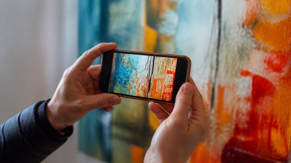 Close-up of hands holding a smartphone displaying high-resolution abstract painting details with vibrant colors and brushwork clearly visible on screen in bright natural lighting