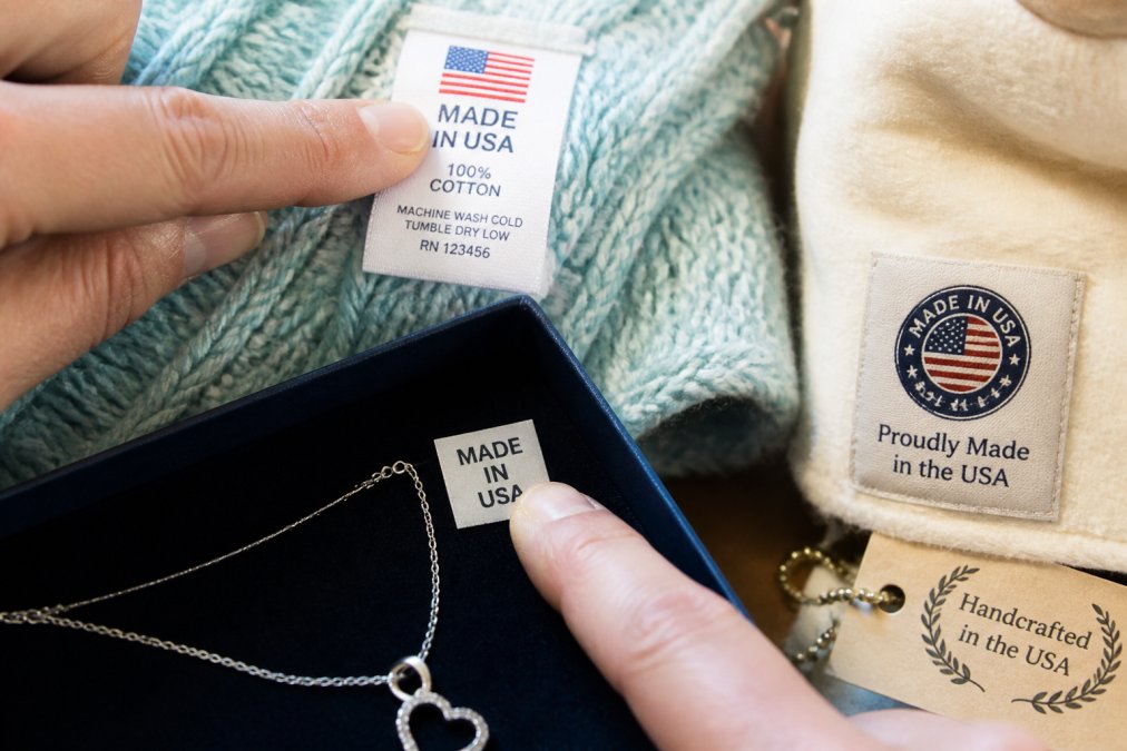 Close-up of product labels and tags showing country of origin markings, certification badges, and manufacturing details on various personalized gift items like jewelry and blankets, with hands pointing to specific label areas