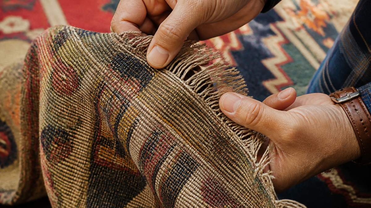 Close-up of a collector's hands examining the backside and fringe of an antique kilim rug, showing weave structure and knot detail under natural light