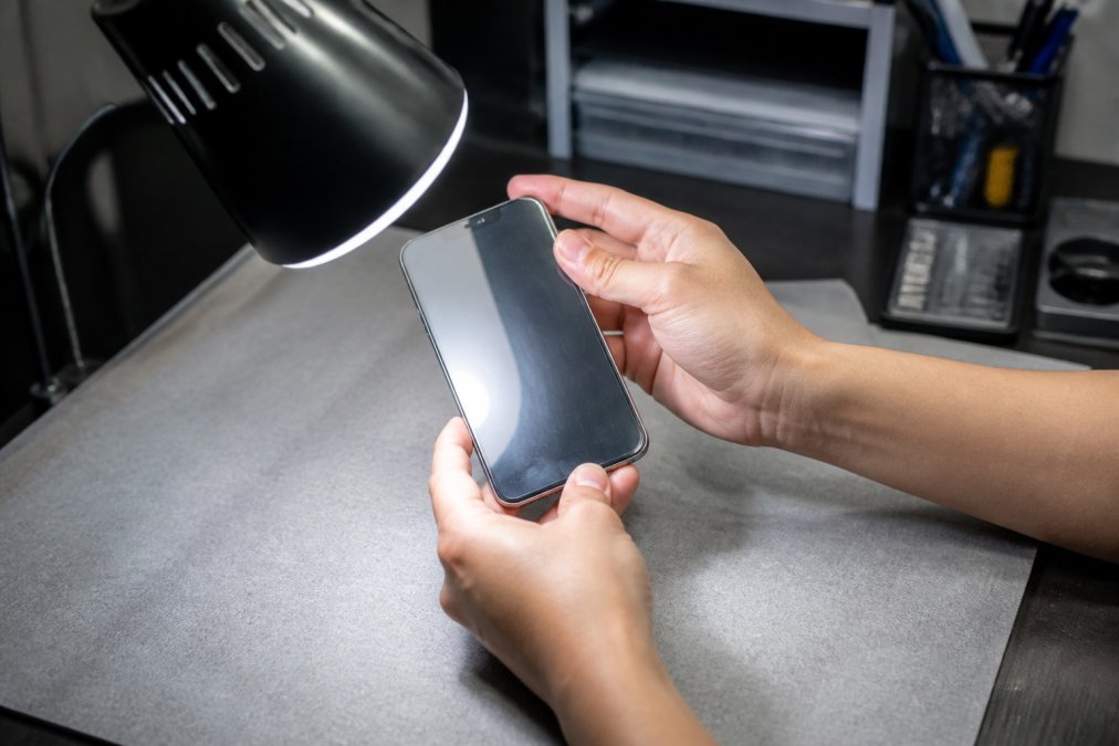 Close-up of a person's hands holding an iPhone under a bright desk lamp, tilting the screen to catch reflections and inspect the glass surface for scratches or uneven edges