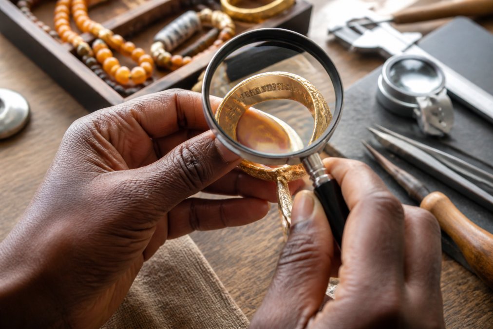 Close-up of hands holding a gold bracelet under bright natural light, using a magnifying glass to inspect hallmarks and maker's marks stamped on the inner metal surface, with jewelry tools visible nearby