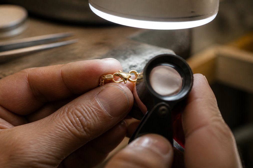 Close-up macro photograph of a jeweller's hands holding a gold necklace clasp under a bright bench lamp, with a loupe and small hallmark stamp visible on the metal surface