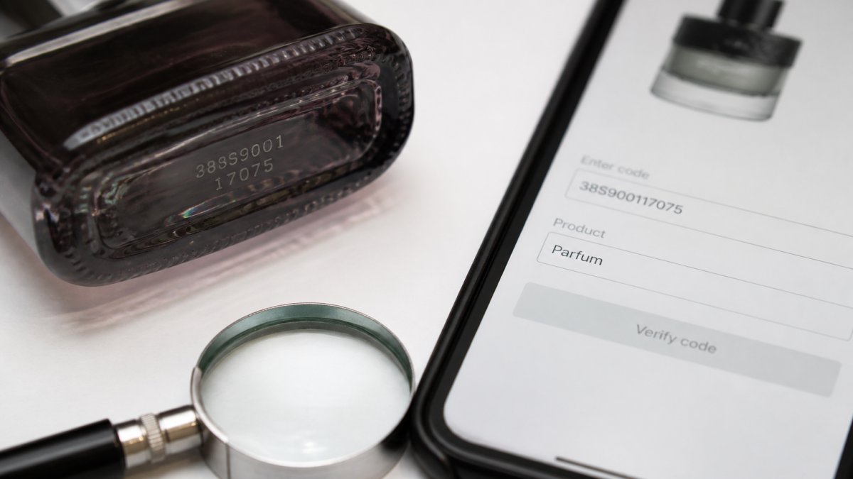 Close-up of a perfume bottle bottom showing batch code numbers next to a smartphone displaying a verification app, with a magnifying glass positioned nearby on white background