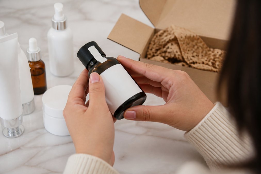 Close-up of a person's hands holding a skincare bottle and examining the label and tamper-evident seal under bright light, with a smartphone nearby displaying a product verification page