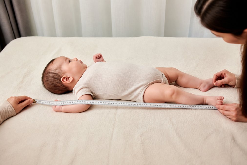 Parent measuring a baby's length lying flat on a soft surface with a cloth tape measure, showing proper technique with the infant straight and relaxed