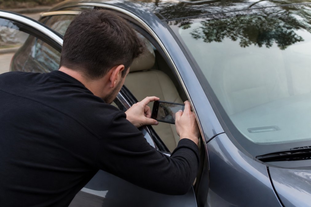 A person standing at the open driver's door of a car, leaning in to look at the VIN plate on the dashboard near the windshield, holding a smartphone to take a photo of it