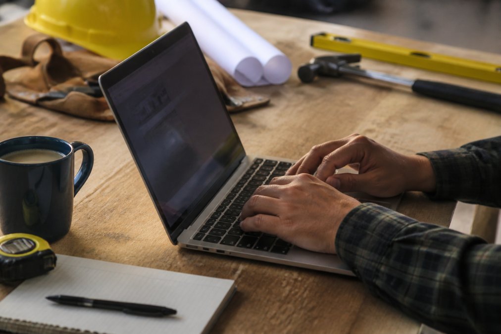 Close-up of hands typing on laptop, displaying a state contractor license lookup website on screen, with a cup of coffee and notepad on desk