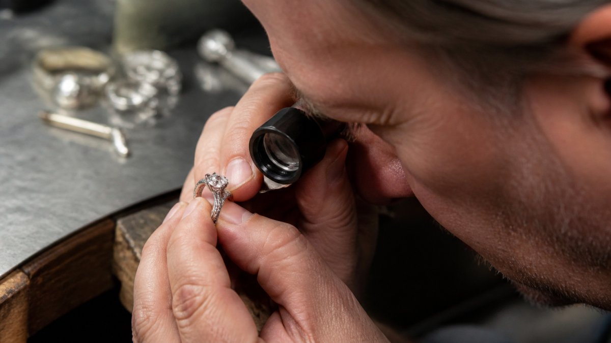Professional jeweler examining a custom diamond ring under a bright magnifying loupe, hands steady and focused on the intricate details of the stone setting and metalwork