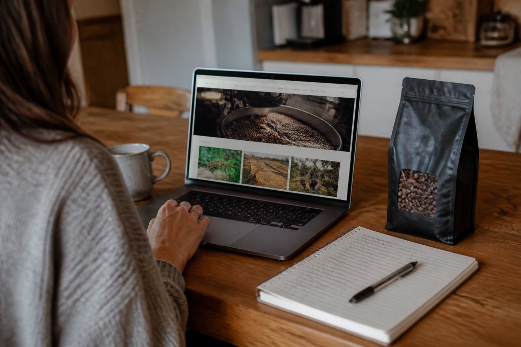 A person sitting at a kitchen table with a laptop open to a coffee roaster's website, a bag of whole bean coffee beside them, and a notebook with handwritten notes about certifications and origin details