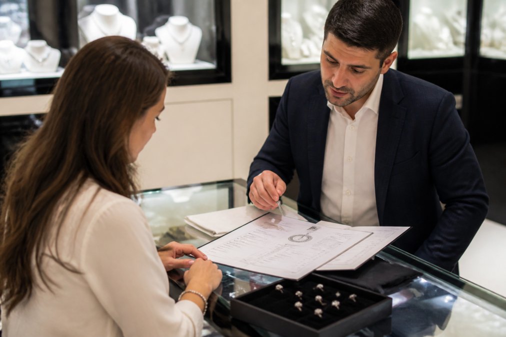 A customer in conversation with a jeweller at a polished counter, reviewing diamond grading certificates and supply chain documentation together, with certified diamonds displayed in soft lighting in the background
