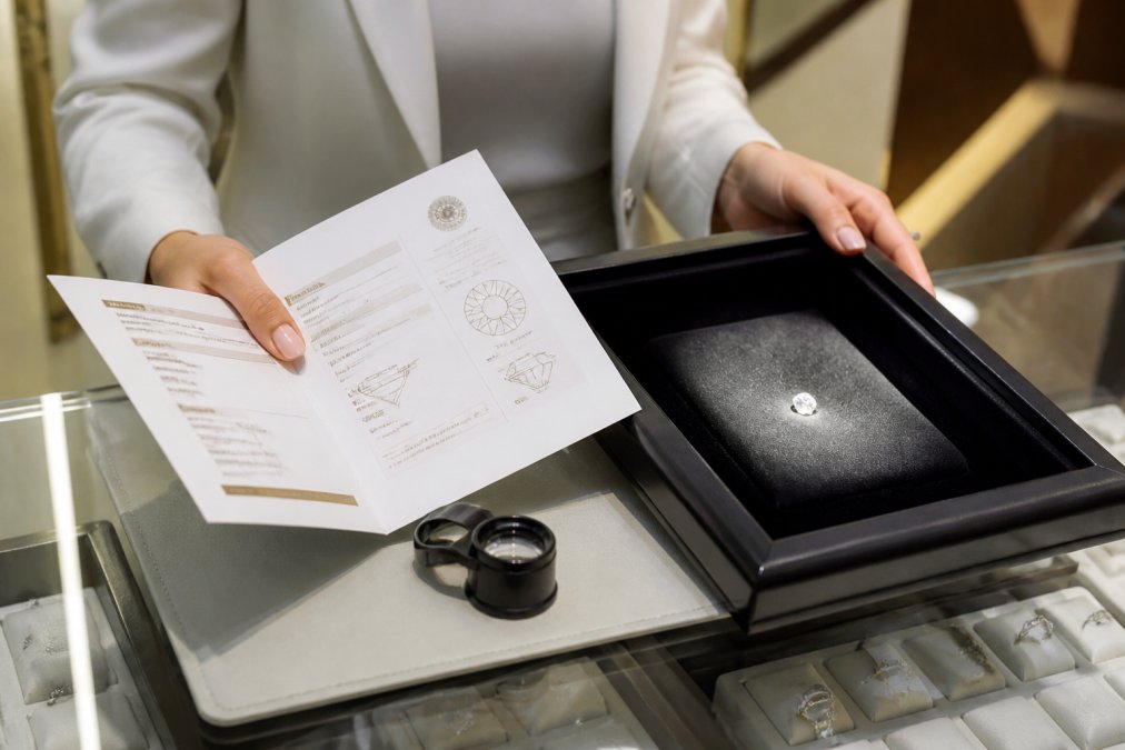 A jeweller's hands presenting a diamond grading report and a small diamond in a lit display tray, with a loupe resting on the counter beside them