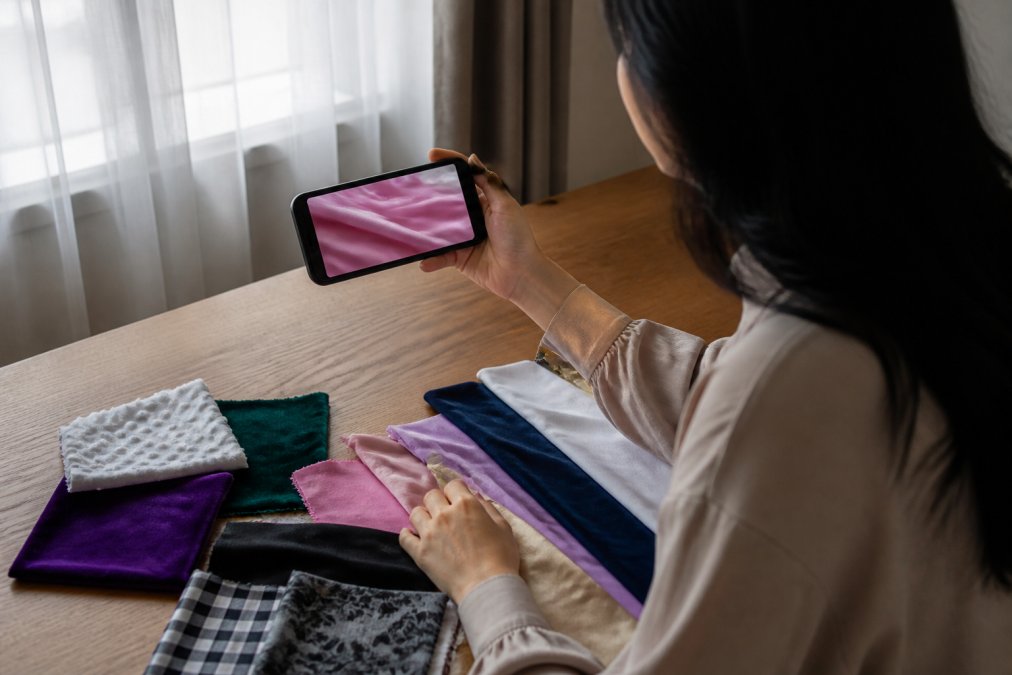 A person sitting at a desk holding a smartphone and watching a slow-motion fabric movement video, with several fabric swatches spread out on the desk beside them in warm natural window light