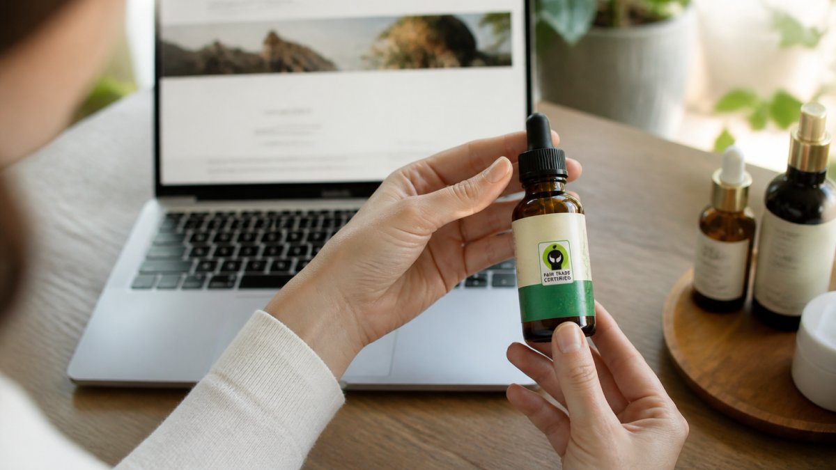 Hands holding a skincare bottle with a fair trade logo to verify fair trade skincare on a wooden desk.