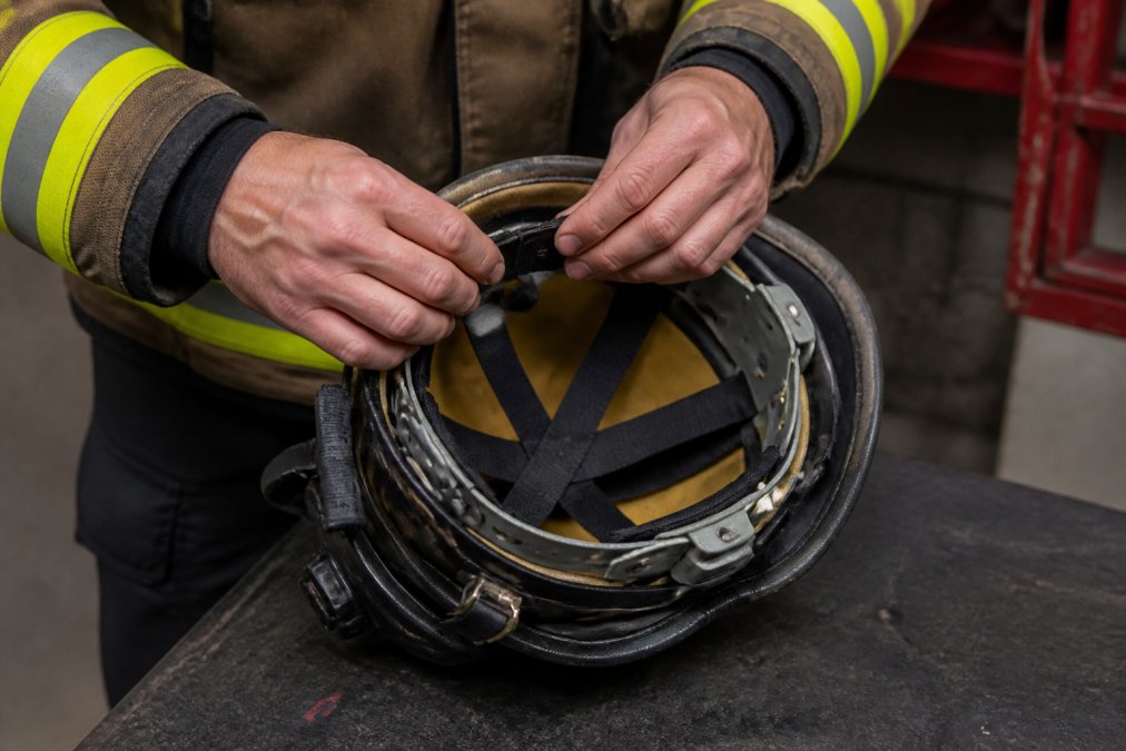 Close-up of a firefighter's hands adjusting the suspension system inside a fire helmet, showing the cradle and elastic bands clearly visible