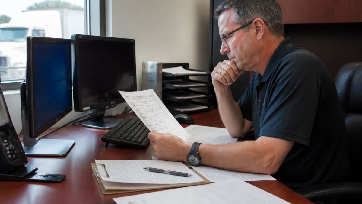 A fleet safety manager reviewing a trucking company's safety rating on a computer screen in an office, with inspection documents and a clipboard on the desk, warm overhead lighting