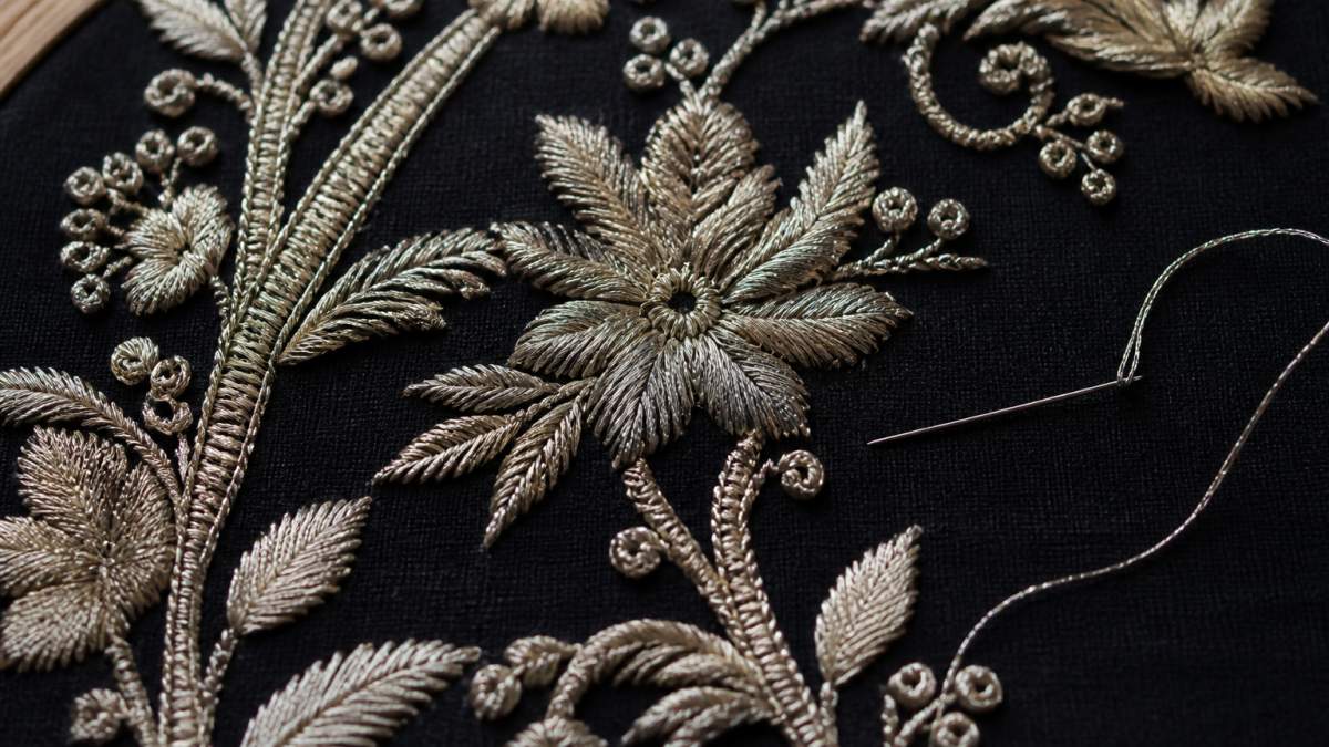 Close-up overhead shot of embroidered fabric showing intricate hand-stitched metallic thread work with visible stitch density, thread tension variations, and detailed needlework against dark fabric background