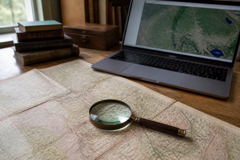 An old historical map spread across a wooden desk beside a laptop showing a modern GIS interface, a magnifying glass resting on the map's faded county lines