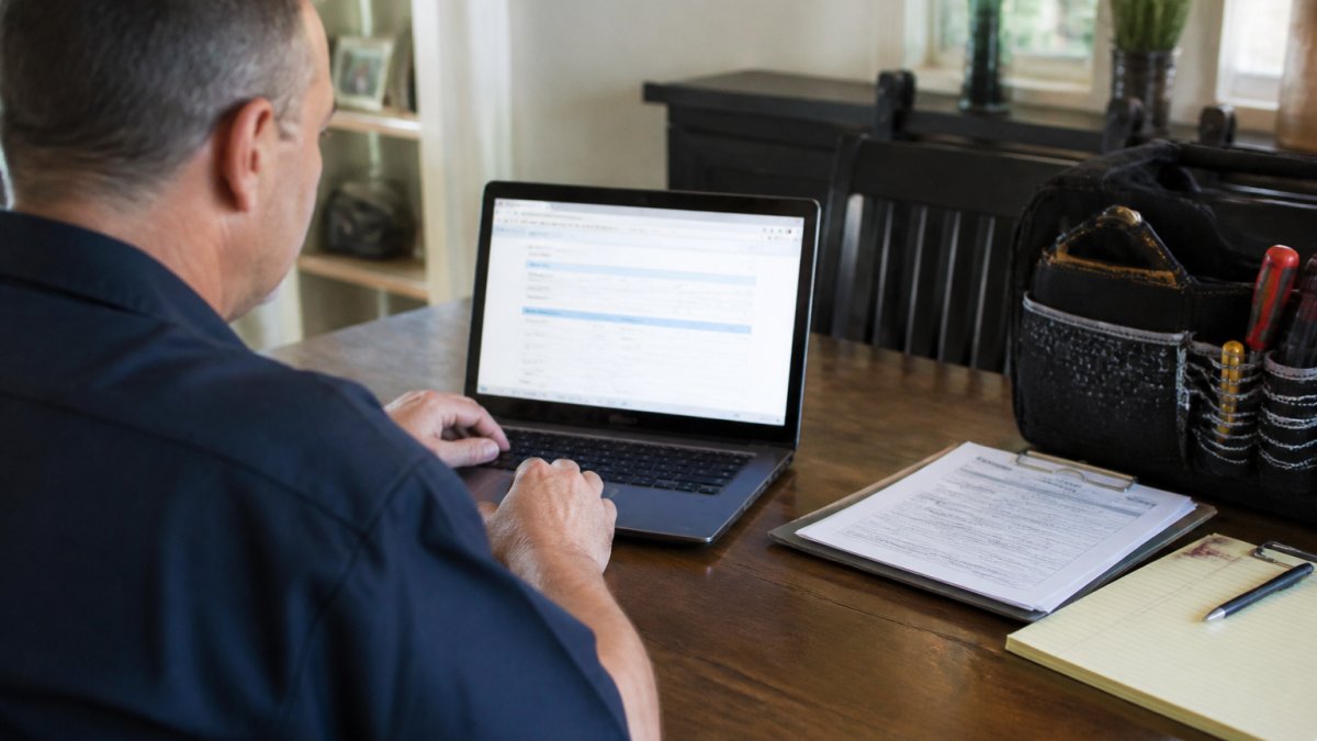 Homeowner sitting at a desk with a laptop, reviewing official state licensing database on screen, with documents and a notepad nearby