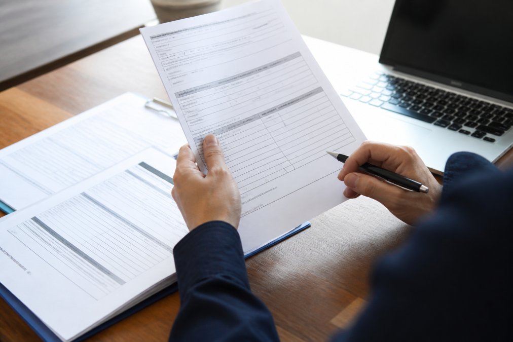 Close-up of hands holding insurance documents and policy paperwork on a desk with a laptop and pen nearby, showing someone reviewing claim details in natural office lighting
