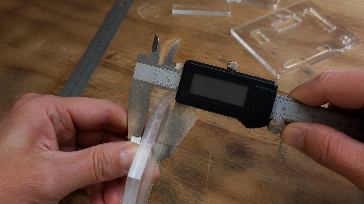 Close-up of hands holding digital calipers against a freshly laser-cut acrylic edge on a workbench, with offcuts and a steel ruler nearby
