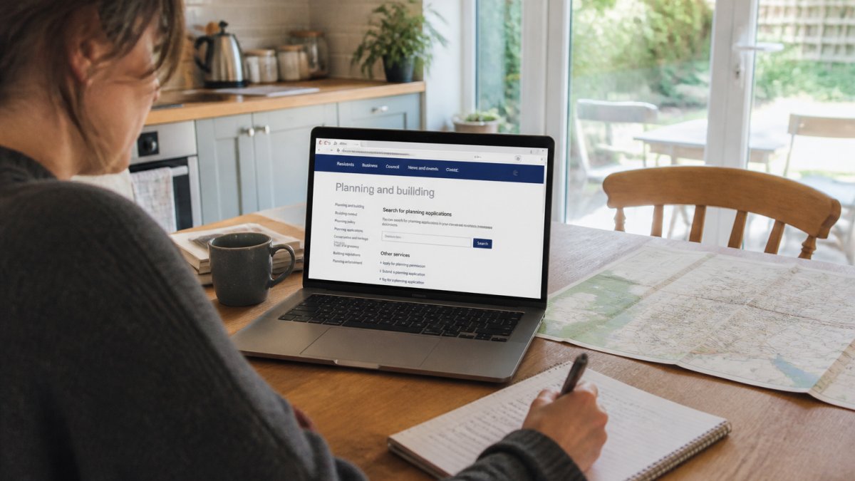 A person at a kitchen table with a laptop open to a council website, a printed map and a notepad with handwritten notes, natural daylight